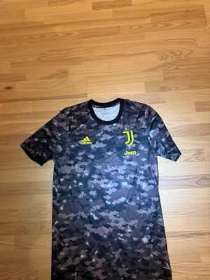 Juventus träningströja Adidas svart - Juventus träningströja från Adidas i svart och grått camomönster med gula detaljer. Tröjan har rund halsringning och korta ärmar. Juventus-logga, Adidas-logga och Jeep-tryck på bröstet. Perfekt för fotbollsträning eller gym.