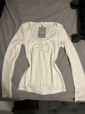 Vit ribbad långärmad topp H&M - Snygg vit ribbad topp från H&M med lång ärm och rundad urringning. Toppen har en smickrande rynkdetalj framtill och är tillverkad i en mjuk viskosblandning. Perfekt att styla med jeans eller kjol för en trendig look.