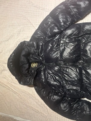 Svart dunjacka från Glorious Gangsta - Snygg svart dunjacka från Glorious Gangsta med glansig finish och huva. Jackan har quiltad design och dragkedja framtill. Perfekt för kalla dagar när du vill hålla stilen och samtidigt vara varm.