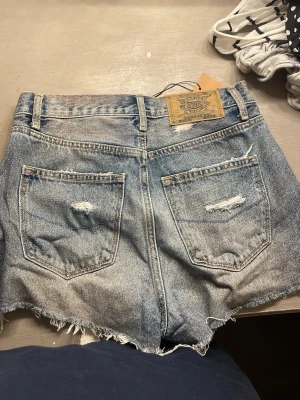 Blå slitna jeansshorts från Levi's - Säljer ett par blå jeansshorts från Levi's med slitna detaljer och fransig kant. Klassisk femficksmodell med hög midja och knappgylf. Perfekta för en avslappnad och trendig look.