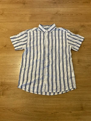 Kortärmad Linneskjorta  - NYPRIS: 300kr    Fräsch vit skjorta med blå ränder från C, tillverkad i en luftig linnemix. Skjortan har korta ärmar, klassisk krage och knäppning framtill. Perfekt för en chill och stilren look.