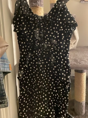 Svart prickig volangklänning - Säljer en svart kortklänning med vita prickar och volangdetaljer. Klänningen har omlottkänsla upptill och är ärmlös med tunna axelband. Perfekt för dig som gillar lekfulla mönster och en feminin look.