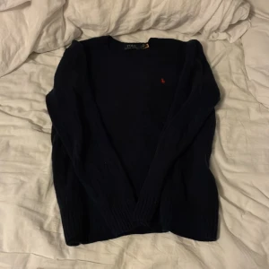 Mörkblå tröja från Polo Ralph Lauren - Mörkblå stickad tröja från Polo Ralph Lauren med rund halsringning och den klassiska röda logotypen broderad på bröstet. Tröjan har långa ärmar och ribbade muddar vid ärmslut och nederkant. Perfekt för en stilren look.
