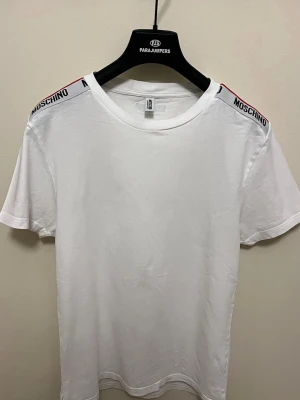 Vit Moschino t-shirt med logoband - Säljer en clean vit t-shirt från Moschino med logoband på axlarna i svart och rött. Klassisk rund halsringning och korta ärmar. Perfekt för dig som gillar stilrena plagg med en twist från ett ikoniskt märke.