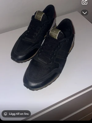 Valentino Rockrunner svarta sneakers - Snygga svarta Valentino Rockrunner sneakers med detaljer i vinrött och beige. Skorna har snörning, rund tå och en mix av skinn och textilmaterial. Klassisk logga på plösen och kontrastfärgad sula för en cool streetwear-look.