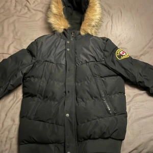 Svart pufferjacka från Zavetti Canada - Säljer en svart pufferjacka från Zavetti Canada med stor huva och fuskpäls. Jackan har dragkedja och knappar framtill, samt en patch på ärmen. Perfekt för kalla dagar och riktigt snygg streetstil.
