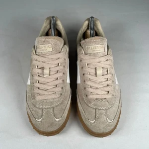 Valentino Upvillage  - Storlek 39 men passar 40. Snygga Valentino Garavani Rockrunner sneakers i beige mocka med vita detaljer och gummisula i brunt. Klassisk låg modell med snörning och ikoniska nitar på sulan. Perfekta för dig som gillar stilrena och exklusiva sneakers med lyxig känsla.