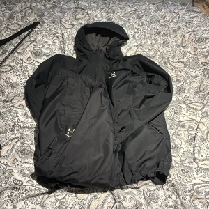 Svart vindjacka från Haglöfs med huva - Säljer en svart vindjacka och byxor set  från Haglöfs med justerbar huva och dragkedja framtill. Jackan har elastiska muddar och dragsko nertill för extra komfort. Materialet är lätt och vattenavvisande, perfekt för blåsiga dagar. Klassisk och stilren design med Haglöfs-logga på bröstet.