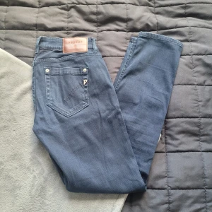 Blå jeans från Dondup, storlek 30 - Snygga blå jeans från Dondup med klassisk femficksdesign och läderpatch bak i midjan. Jeansen har raka ben och silverfärgade nitar. Perfekta för dig som gillar en stilren look med italiensk touch.