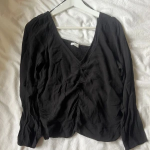 Svart v-ringad blus med rynk framtill - Svart blus  med v-ringning och rynkdetalj framtill. Långa ärmar och smockad rygg för en skön passform. Tunn och luftig i materialet, perfekt till jeans eller kjol. Klassisk och stilren modell.