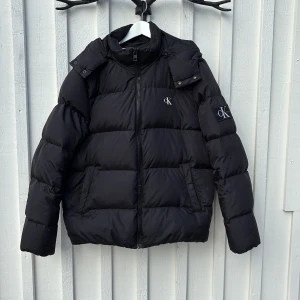 Svart pufferjacka från Calvin Klein stl XL - Säljer en snygg svart pufferjacka från Calvin Klein Jeans. Jackan har en dragkedja framtill och en broderad logga på ärmen. Perfekt för kyliga dagar med sin vadderade design och huva. Säljes i mycket bra begagnat skick. 8/10 