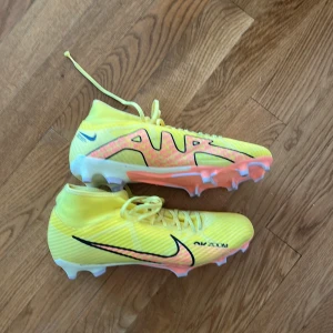Nike Air Zoom gula fotbollsskor - Säljer ett par Nike Air Zoom fotbollsskor i en riktigt fräsch gul färg med svarta och rosa detaljer. Skorna har snörning, högre skaft och vit sula med dobbar. Perfekta för dig som vill sticka ut på planen och ha grymt grepp. Snygg design och lätt material.