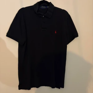 Svart pikétröja från Polo Ralph Lauren - Klassisk svart pikétröja från Polo Ralph Lauren med korta ärmar och krage. Tröjan har den ikoniska röda loggan broderad på bröstet och knäppning framtill. Perfekt basic med snygg passform och tidlös stil.