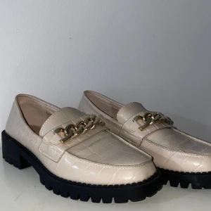 Beige loafers med kedjedetalj Primark - Snygga beige loafers från Primark i storlek 37 med croco-mönstrad yta och chunky svart sula. Skorna har en fet guldfärgad kedjedetalj framtill som ger en cool touch. Perfekta för dig som vill sticka ut med stilrena men trendiga skor.