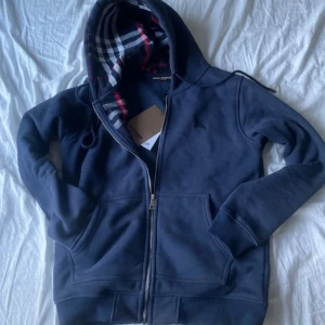 Marinblå hoodie från Burberry - Snygg marinblå hoodie från Burberry med klassiskt rutigt foder i huvan. Tröjan har dragkedja framtill, broderad logga på bröstet och stora fickor.