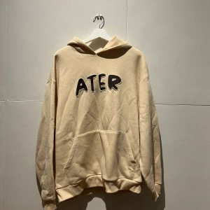 Beige hoodie med svart text ATER - Säljer en beige hoodie från ATER med stor svart text broderad på bröstet. Tröjan har huva, känguruficka och långa ärmar med ribbade muddar. Perfekt för dig som gillar streetwear och vill ha en chill look.
