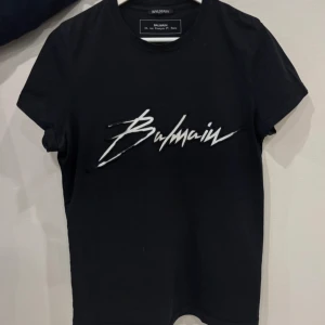 Svart Balmain t-shirt med broderad logga - Svart t-shirt från Balmain med vit broderad logotyp framtill i coolt, handskrivet typsnitt. Klassisk rund halsringning och korta ärmar. Perfekt för dig som gillar exklusiva streetwear-vibbar.