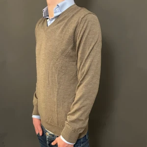 Beige v-ringad stickad tröja - Snygg beige stickad tröja med v-ringning och långa ärmar. Perfekt att ha över en skjorta för en clean och chill look. Tröjan har ribbade muddar vid ärmslut och nederkant, vilket ger en schysst passform. Passar dig som gillar klassisk stil med en modern touch.