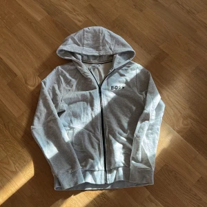 Grå hoodie från BOSS med dragkedja - Snygg ljusgrå hoodie från BOSS med svart dragkedja och huva. Tröjan har BOSS-logga på bröstet och är tillverkad i mjuk bomullsmix. Perfekt för en chill och avslappnad stil. Fickor på sidorna och ribbade muddar vid ärmslut och nederkant.