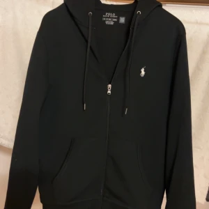 Svart hoodie från Polo Ralph Lauren - Svart hoodie från Polo Ralph Lauren med dragkedja, huva med snörning. Köpt för 2199 för runt 2-3 månader sen men den var lite stor för mig vilket har lett till knappt någon användning. Kvitto och påse kommer med på köp. Strl XS men skulle säga att den är som en S i strl