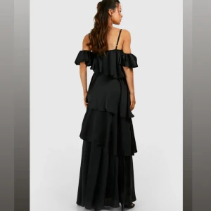 Svart volangklänning  - Säljer en så fin svart långklänning med tunna axelband och offshoulder-volanger. Klänningen har flera lager med volanger och en v-ringad urringning. Helt ny och aldrig använd 