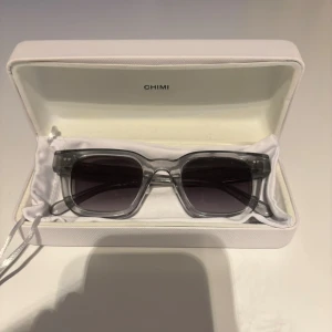CHIMI solglasögon i grå - Snygga solglasögon från CHIMI med transparent grå båge och mörka rektangulära linser. Kommer i original vit hård ask med logga. Perfekt accessoar för att lyfta din stil och skydda ögonen mot solen.