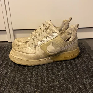 Nike Air Force 1 vita sneakers - Nike Air Force 1 sneakers i beige med klassisk rund tå och snörning. Skorna har en platt sula och är tillverkade i en mix av skinn och syntetmaterial. Ikonisk Swoosh-logga på sidorna och perforerade detaljer på tån för extra ventilation. Skolåda medföljer