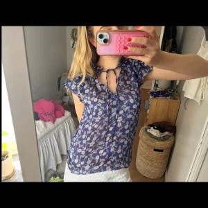 Blommig blå blus med knytband - Söt blommig blus i mörkblått med ljusa blommor. Tunn och luftig med korta volangärmar och v-ringning med knytband vid halsen. Perfekt för varma dagar och ger en romantisk vibe till outfiten.