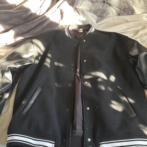 Svart bomberjacka från H&M Divided XS - Svart bomberjacka från H&M Divided i storlek XS. Jackan har skinnliknande ärmar, ribbad krage och muddar med vita ränder. Framsidan har knappar och två fickor med skinnliknande detaljer.