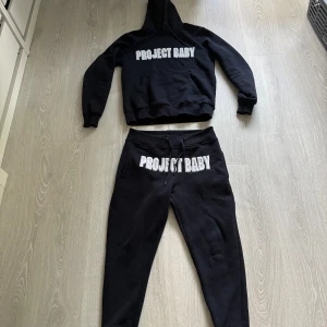 Svarta mjukisbyxor Project Baby - Svarta mjukisbyxor från Project Baby med broderad text i vitt framtill. Byxorna har dragsko i midjan och ribbade muddar vid bensluten. Perfekta för en chill och avslappnad stil.