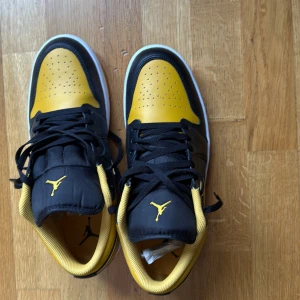Nike Air Jordan 1 Low svart/gul - Nike Air Jordan 1 Low sneakers i svart och gul med klassisk Jumpman-logga på plösen och insidan. Skorna har snörning, perforerad tå och vit sula. Låg modell med rund tå och platt sula. Materialet är skinn och syntet, vilket ger en snygg och sportig vibe.