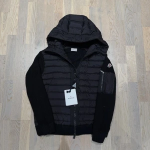 Svart cardigan jacka från Moncler - Svart cardigan jacka från Moncler med quiltad pufferfront och stickade ärmar. Jackan har huva, dragkedja och en ficka med Moncler-logga på ärmen. Perfekt för dig som vill ha en snygg och modern look med både komfort och stil till vintern. Tveka inte på att höra av er vid funderingar!