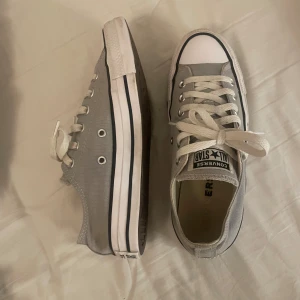 Grå Converse All Star sneakers - Säljer ett par klassiska Converse All Star sneakers i grått tyg med vita gummisulor och vita snören. Skorna har rund tå, lågt skaft och tydliga logodetaljer på plösen. Dom är absolut använda, men är fortfarande bra skick förutom att hälen är uppsliten som på bilden.  