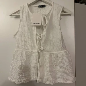 Volangväst med snörning från Ambar - Säljer en söt volangväst från Ambar i både vit och svart. Västen har en strukturerad yta, knytband framtill och volangdetaljer nedtill. Perfekt att styla över en t-shirt eller klänning för en trendig look. Finns i två färger – välj din favorit!