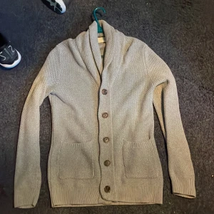 Grå  kofta från Selected Homme - Säljer en grå stickad kofta från Selected Homme med grov struktur, sjalkrage och stora bruna knappar. Koftan har två framfickor och ribbade muddar vid ärmslut och nederkant. Perfekt för dig som gillar chill och stilren look.