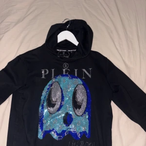  Philipp Plein hoodie  - Cool svart hoodie från Philipp Plein med en stor glittrig blå och silverfärgad Pacman-inspirerad design framtill. Tröjan har huva och är långärmad, perfekt för dig som vill sticka ut med en unik och lyxig streetstyle.