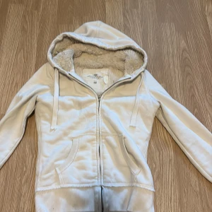 Beige hoodie med fuskpäls från H&M - Mysig beige hoodie från H&M L.O.G.G. med fluffigt fuskpälsfoder på insidan. Tröjan har huva med snörning, dragkedja framtill och två fickor. Perfekt för kyliga dagar när du vill vara både varm och snygg.