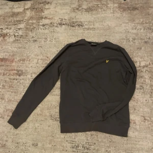 Mörkgrå sweatshirt från Lyle & Scott - Mörkgrå sweatshirt från Lyle & Scott med klassisk gul broderad logga på bröstet. Tröjan har rund halsringning, långa ärmar och ribbade muddar. Perfekt för en chill och stilren look. Den är oanvänd och helt ny.