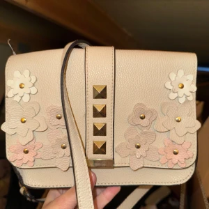 Beige axelväska med blommor och nitar - Superfin beige axelväska i skinn med 3D-blommor ett rymligt fack med dragkedja inuti. Perfekt för dig som gillar detaljer och vill sticka ut lite extra.