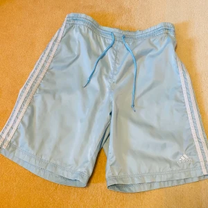 Ljusblå Adidas shorts med snörning - Snygga ljusblå shorts från Adidas med klassiska vita ränder längs sidorna och broderad logga. De har snörning och är tillverkade i lätt polyester med meshfoder. Skönt glansigt material!