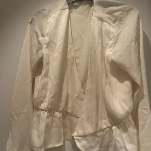 Vit omlottblus från BikBok - Säljer en stilren vit omlottblus från BikBok i storlek M. Blusen har långa ärmar, knytband i midjan och lätt glansigt material som ger en lyxig känsla. Perfekt för dig som gillar en feminin och trendig look.