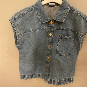 Jeansväst från Name It med guldknappar - Snygg jeansväst från Name It i klassisk blå denim. Västen har krage, en bröstficka och guldiga knappar framtill. Avslutet nertill är lätt rynkat för en cool look. Perfekt att slänga över en t-shirt för en avslappnad stil. Pris går att diskutera 
