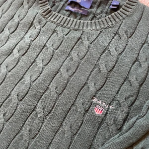 Grön kabelstickad tröja från GANT - Mörkgrön kabelstickad tröja från GANT i 100% bomull. Klassisk rund halsringning, ribbade muddar och broderad GANT-logga på bröstet. Perfekt för dig som gillar preppy stil och vill ha en tidlös tröja med snygga detaljer.