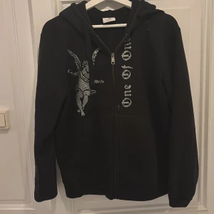 Svart Rhinestone hoodie - One of One - Svart hoodie från One of One i storlek S med dragkedja och huva. Framsidan har en cool ängel och texten 'One of One' i strasstenar. Perfekt för dig som gillar streetstyle och vill sticka ut lite extra.