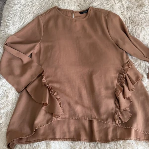Beige volangsett från SHEIN XS - Supersöt brun sett från SHEIN i storlek XS. Blusen har långa ärmar och snygga volangdetaljer längs sidorna. Den är tillverkad i ett mjukt, lätt material och har en avslappnad passform med rund halsringning. Perfekt för dig som gillar en stilren men ändå lekfull look.