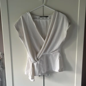 Vit omlottopp från Zara - Säljer en stilren vit omlottopp från Zara med knyt i midjan. Toppen har v-ringning och är ärmlös, vilket ger en fräsch och modern look. Perfekt att matcha med jeans eller kjol för en clean stil. Ej använt 