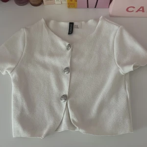 Vit kortärmad topp med knappar H&M - Superfin vit kortärmad topp från H&M Divided med tre silvriga knappar framtill. Modellen är croppad och har en lätt struktur i tyget. Perfekt för dig som gillar en enkel men ändå snygg look.