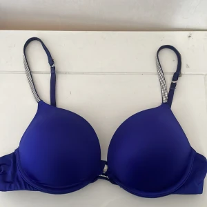 Blå push-up bh med strassband - Snygg blå push-up korsett med glansigt material och vadderade kupor. Axelbanden är dekorerade med strassdetaljer som ger extra bling. Perfekt för dig som vill ha en cool och glam look under kläderna. Justerbara band för bästa passform.