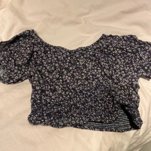Svart blommig croppad blus - Säljer en svart blommig blus med lila och gröna små blommor. Toppen är croppad med smockad midja och har korta puffärmar. Perfekt för dig som gillar romantisk stil och vill ha något som sticker ut lite extra.