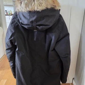 Svart parkas från Canada Goose  - Svart parkas från Canada Goose med stor huva och beige pälsdetalj. Jackan har klassisk logga på ärmen, dragkedja och knappar framtill samt flera fickor. Perfekt för kalla dagar och har en riktigt snygg och tidlös look.